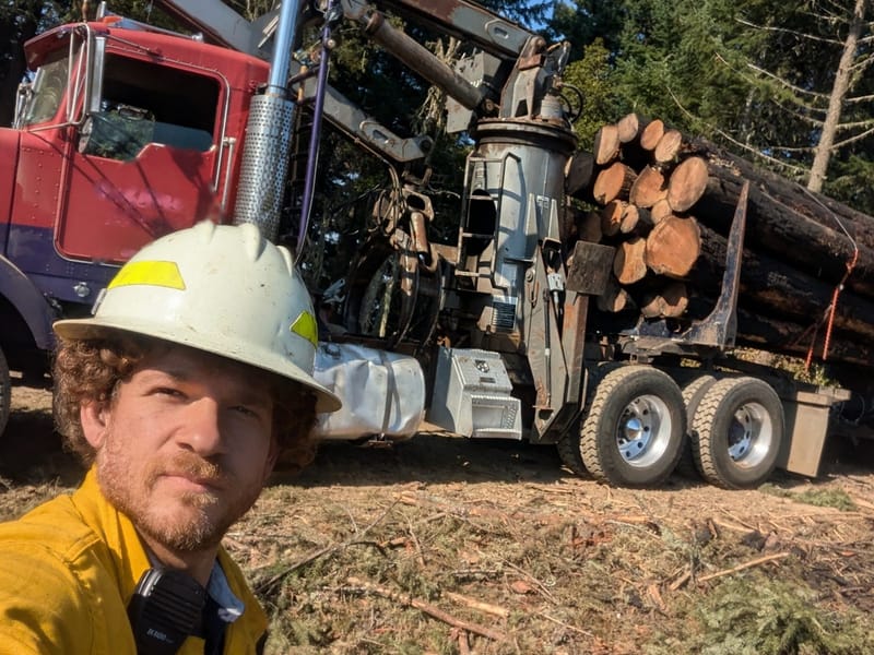Omer in a yellow fire shirt and white hard hat, beside a red self-loader log truck loaded with logs in a cleared stretch of forest.
